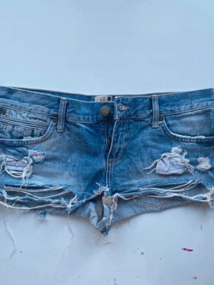 Blå slitna jeansshorts med fransar - Säljer ett par blå jeansshorts med råa, slitna detaljer och fransar längs kanterna. Shortsen har klassisk femficksdesign, bälteshällor och knappgylf. Perfekta för en avslappnad och edgy stil. Ca 39cm i midjemått rakt över