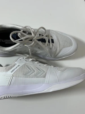 Vita Hummel Power Play Pro sneakers - Säljer ett par vita Hummel Power Play Pro sneakers med klassisk låg profil och snörning. Skorna har ventilerande meshpaneler, syntetiska detaljer och märkets ikoniska pilar på sidan. Perfekta för sport eller träning.OBS! Skorna har ett litet hål.