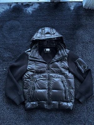 C.P. Company cardigan - Snygg svart pufferjacka från C.P. Company med stickade ärmar och huva. Jackan har två stora framfickor med dragkedja och en ikonisk detaljficka på ärmen. Helt ny så skick 10/10, storlek M/L, pris går att diskutera✅🐼