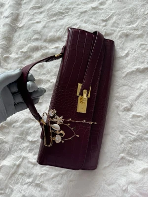 Vinröd axelremsväska med krokoprägling - Snygg vinröd handväska i avlång modell med krokopräglat mönster och guldfärgade detaljer. Väskan har ett dekorativt hängsmycke med snäckor och stjärnor på axelremmen samt ett elegant guldfärgat lås framtill. Perfekt accessoar för att lyfta din outfit.