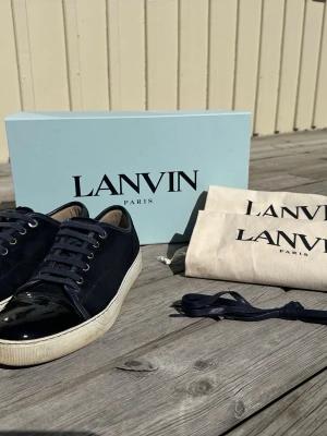 Mörkblå Lanvin sneakers med lackdetalj - Snygga mörkblå sneakers från Lanvin med klassisk låg profil. Skorna har ovandel i mocka och tåparti i blankt lack, samt vita sulor. Kommer med extra skosnören och dustbags. Perfekta för dig som gillar stilrena och exklusiva sneakers. Kan maila kvitto från care of Carl vid köp.
