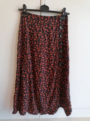 Blommig midikjol med slits - En rödblommig svart midikjol med dekorativa knappar längs sidan ovanför slitsen. Kjolen har en lätt utsvängd form och är perfekt för dig som gillar en feminin stil. Gjord i viskos som känns mjukt och följsamt (tyvärr klippt bort lappen som skavde). 