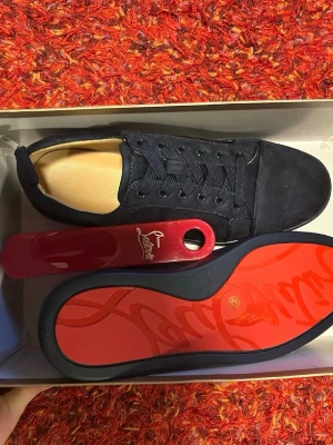 Svarta Louboutin sneakers med röd sula - Snygga svarta sneakers från Christian Louboutin i mocka med ikonisk röd sula och svarta skosnören. Låg modell med diskret logga på sidan och guldfärgat innerfoder. Perfekta för dig som vill sticka ut med exklusiva detaljer.