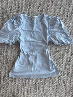 Ljusblå topp med puffärmar - Trendig ljusblå topp med stora puffärmar och rund halsringning. Ärmarna är i ett tunnare, lite glansigt tyg. Perfekt för dig som vill ha en söt och stilren look.