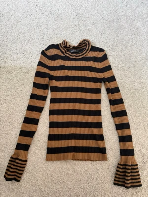 Randig stickad tröja Gina Tricot - Stickad tröja från Gina Tricot med svarta och bruna ränder. Tröjan har lång ärm, volangdetaljer vid halsringning och ärmslut samt ribbad struktur. Perfekt för dig som gillar trendiga detaljer och snygga färgkombinationer.