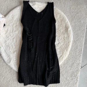 Svart stickad ärmlös klänning (S) - Svart stickad klänning med v-ringning och ärmlös design. Klänningen har ett ribbat parti nedtill och små glittriga detaljer i tyget. Snygga slitningar framtill ger en edgy look. Perfekt för dig som gillar en unik stil.