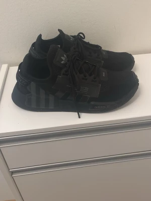 Svarta Adidas NMD sneakers - Snygga helsvarta Adidas NMD sneakers med stickad ovandel och klassiska tre ränder på sidan. Skorna har svarta snören, diskreta detaljer och en bekväm sula. Perfekta för dig som gillar stilrena och sportiga sneakers med modern design.