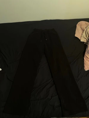 Svarta vida mjukisbyxor - Säljer ett par svarta mjukisbyxor med breda ben och resår i midjan samt dragsko. Perfekta för chill dagar hemma eller när du vill ha en avslappnad stil. Materialet känns mjukt och bekvämt mot huden.