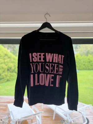 Svart sweatshirt med rosa text - Säljer en svart sweatshirt med stort rosa tryck på framsidan där det står 'I SEE WHAT YOU SEE AND I LOVE IT'. Tröjan har rund halsringning och långa ärmar. Perfekt för dig som gillar statement-plagg och vill sticka ut lite extra.
