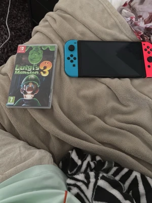 Nintendo Switch med blå och röd Joy-Con - Nintendo Switch spelkonsol med avtagbara Joy-Con kontroller i neonblått och neonrött. Konsolen har en svart kropp och stor skärm. Perfekt för gaming både hemma och på språng. Spelboxen Luigi's Mansion 3 syns också på bilden. Super mario finns också!!!!!!!!!!!!! Den är helt ny