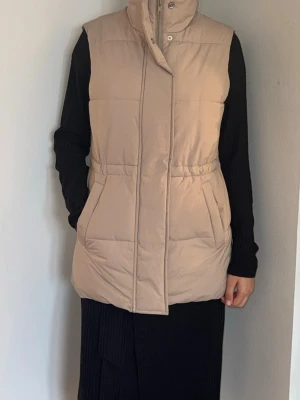 Beige lång dunväst med hög krage - Stilren beige dunväst med hög krage och dold dragkedja framtill. Västen har två stora fickor och markerad midja för en snygg siluett. Perfekt att slänga över en hoodie eller stickad tröja för extra värme. Lång modell som ger en trendig look.