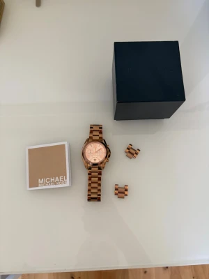 Rosé klocka från Michael Kors - Snygg rose klocka från Michael Kors med länkarmband i metall och rund urtavla. Klockan har kronograf och detaljer i roseguld. Nyskick. Ingår även 2 bitar för att göra armbandet större vid behov. 