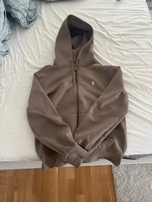 Brun hoodie med dragkedja från Polo Ralph Lauren - Säljer en brun hoodie från Polo Ralph Lauren med dragkedja framtill och klassisk vit logga på bröstet. Hoodien har huva med dragsko och två fickor framtill. Tillverkad i mjukt bomullsmaterial som är skönt att ha på sig.