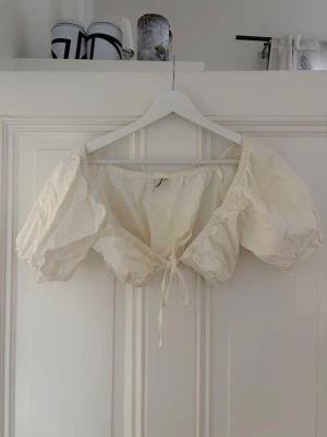 Vit omlott bolerotopp med puffärm - Supersöt vit bolerotopp med puffiga korta ärmar och omlottknytning framtill. Toppen har en croppad passform och är gjord i ett lätt bomullstyg. Perfekt för att styla över linnen eller klänningar för en trendig look.
