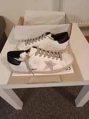White Golden Goose sneakers - Säljer ett par vita sneakers från Golden Goose med svarta detaljer och beige sula. Skorna har en klassisk låg siluett och är tillverkade i läder med perforerade hål på sidan. Snygg och trendig design som sticker ut.