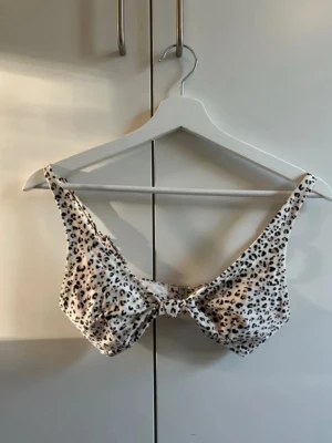 Leopardmönstrad bikiniöverdel med knyt - Snygg bikiniöverdel i vitt med leopardmönster och knyt framtill. Modellen har breda axelband och är tillverkad i ett mjukt syntetmaterial som torkar snabbt. Perfekt för strandhäng eller pooldagar.