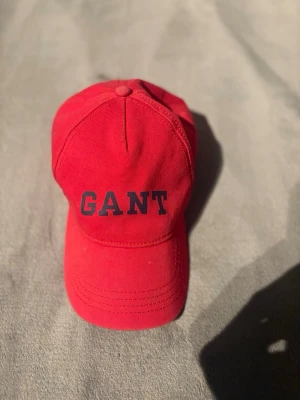 Gant keps  - Snygg röd keps från GANT med klassisk böjd skärm och justerbar rem bak. Kepsen har GANT-loggan broderad i mörkblått framtill och är tillverkad i bomullstyg. Perfekt accessoar för en sportig och avslappnad stil.