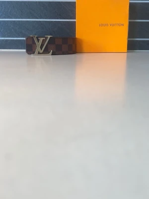 Brunt rutigt bälte i skinn - Kommer med box, Snyggt brunt bälte  med klassiskt rutigt mönster och stor logga i guld som spänne. Tillverkat i skinn och perfekt för att lyfta vilken outfit som helst. Ett måste för dig som gillar exklusiva accessoarer.