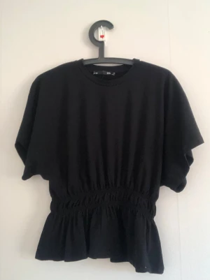Svart topp med resår och volang - Snygg svart topp med rund hals, korta ärmar och resår i midjan som ger en markerad siluett. Volang nedtill för en extra touch. 🤩