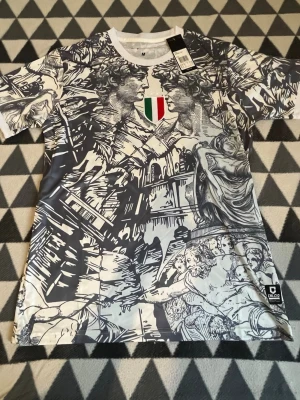 Grafisk t-shirt med italienskt motiv - Unik t-shirt med svartvitt grafiskt tryck av klassiska italienska statyer och byggnader. Framtill finns en liten italiensk flagga på bröstet. T-shirten har rund hals och korta ärmar, perfekt för dig som gillar konstnärliga och iögonfallande plagg.