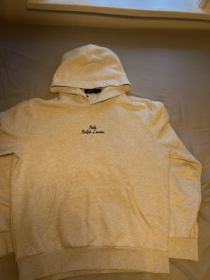 Beige hoodie från Polo Ralph Lauren - Säljer en beige hoodie från Polo Ralph Lauren med broderad logga på bröstet. Hoodien har en klassisk känguruficka framtill och mjukt material som känns skönt mot huden. Perfekt för dig som gillar stilrena och bekväma plagg.