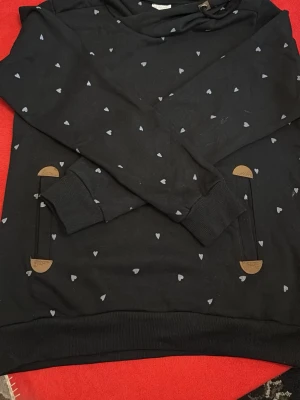 Svart hoodie med små hjärtan Infinity - Svart hoodie från Infinity med små vita hjärtan över hela tyget. Hoodien har en huva med snörning och detaljer i brunt konstläder vid fickorna och huvan. Muddar vid ärmslut och nederkant för extra komfort. Perfekt för en avslappnad och trendig look.