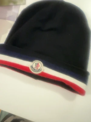 Svart Moncler mössa med logga - Snygg svart mössa från Moncler med klassisk Moncler-logga framtill. Mössan har en uppvikt kant med ränder i blått, vitt och rött som ger en sportig vibe. Tillverkad i mjukt material som känns skönt mot huden. Perfekt accessoar för kalla dagar.