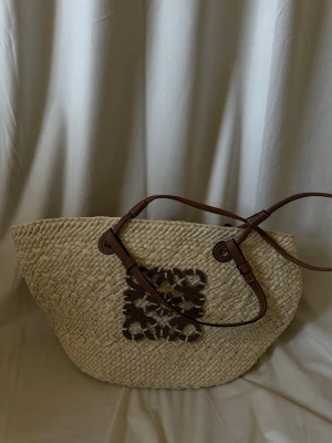 Beige stråväska med bruna detaljer - Snygg shoppingväska i beige stråmaterial med bruna läderhandtag och dekorativt brunt mönster framtill. Väskan har en rymlig form och passar perfekt för dig som vill ha en trendig och praktisk accessoar till vardagen.