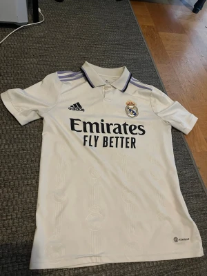 Vit Real Madrid matchtröja Adidas - Säljer en vit Real Madrid matchtröja från Adidas med klubbmärke och Adidas-logga på bröstet. Tröjan har svarta detaljer, krage med lila och svart kant samt trycket 'Emirates Fly Better' på framsidan. Tillverkad i ett lätt och ventilerande material.