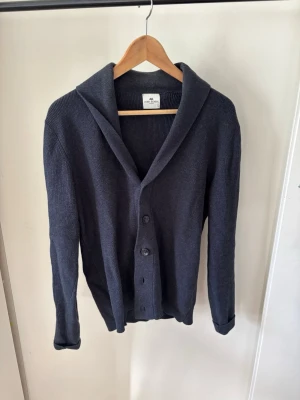John Henric Cardigan - Mysig mörkblå stickad kofta med bred krage och tre knappar framtill. Koftan har lång ärm och ribbade muddar vid ärmslut och nederkant. Perfekt för lager på lager och enkel att matcha med olika outfits.