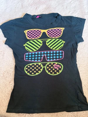 Svart t-shirt med färgglada solglasögon - Cool svart t-shirt från Punkyfish med tryck av fyra färgglada solglasögon i olika mönster. T-shirten står strl L men de rimligare att det är M passform eller mindre strl!! 