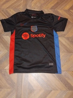 Svart FC Barcelona matchtröja Nike - Snygg svart FC Barcelona matchtröja från Nike med röda och blå detaljer på sidorna. Tröjan har klubbmärke, Nike-logga och Spotify som sponsor framtill. Tillverkad i Dri-FIT material som andas, perfekt för träning eller match.