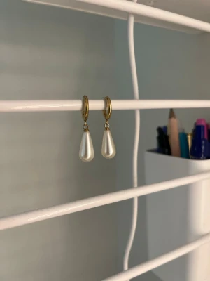 Guldiga örhängen med droppformade pärlor - Säljer ett par stilrena örhängen med guldfärgade ringar och droppformade vita pärlor. Perfekta för dig som gillar klassiska accessoarer med en modern twist. Materialet ser ut att vara metall och pärlimitation. Handgjort och rostfritt stål