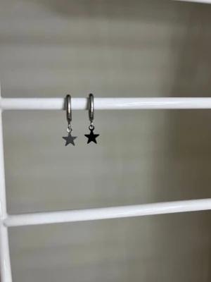 Silvriga örhängen med stjärnhängen - Säljer ett par stilrena silvriga örhängen med små stjärnformade hängen. Örhängena har en enkel ring som bas och stjärnorna hänger fritt. Perfekta för dig som gillar minimalistiska smycken med en twist. Handgjort och rostfritt stål