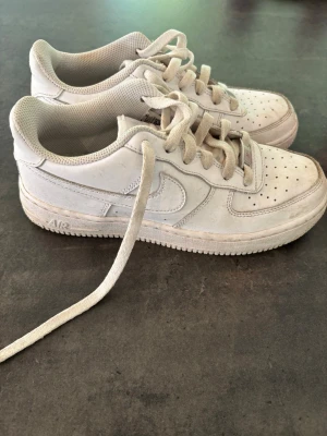 Vita Nike Air Force 1 sneakers - Klassiska vita Nike Air Force 1 sneakers med lågt skaft och perforerad tå. Skorna har en chunky sula med AIR-detalj på sidan och är tillverkade i läder med textilfoder. Perfekta för en clean och stilren look.