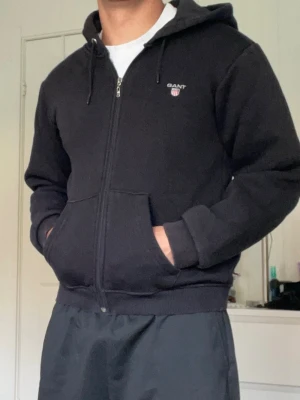 Svart zip hoodie från GANT - Klassisk svart hoodie från GANT med dragkedja, huva med dragsko och två stora fickor framtill. Diskret GANT-logga broderad på bröstet. Perfekt för en avslappnad och sportig stil.