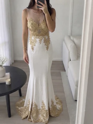 Fest klänning  - Elegant vit långklänning med detaljerad guldspets upptill och längs fållen. Klänningen har ärmlös design, mesh upptill och figurnära passform som ger en lyxig känsla. Perfekt för bal eller festliga tillfällen.