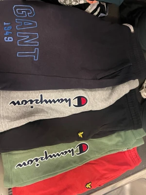 Mjukisbyxor från Gant, Champion & Lyle & Scott - Flera par mjukisbyxor i olika färger och märken: mörkblå från Gant med blå logga, grå och gröna från Champion med broderad logga längs benet, samt svarta och röda från Lyle & Scott med gul brodyr. Perfekta för chill eller träning.