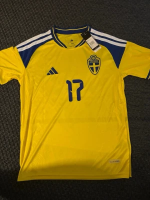 Svensk landslagströja Adidas gul Gyökeres S - Gul svensk landslagströja från Adidas med blå detaljer, nummer 17 på bröstet och det klassiska Sverige-emblemet. Tröjan har rund hals, blå ränder på axlarna och är tillverkad i ett lätt, tekniskt material. Perfekt för fotboll eller att bära på matchdag. S