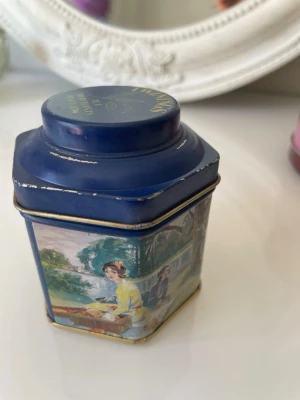 Blå vintage teburk från Twinings - Säljer en blå fyrkantig plåtburk från Twinings med motiv av människor i parkmiljö. Burken har guldfärgade detaljer och texten 'Tea Merchants Since 1706' på locket. Perfekt som dekoration eller förvaring av småsaker.