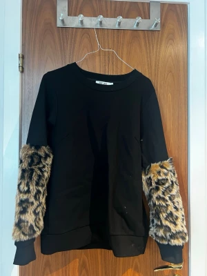 Svart sweatshirt med fuskpälsärmar - Cool svart sweatshirt från DRY LAKE med unika ärmar i leopardmönstrad fuskpäls. Tröjan har rund halsringning och muddar vid ärmslut och nederkant. Perfekt statement-plagg för dig som vill sticka ut med din stil.