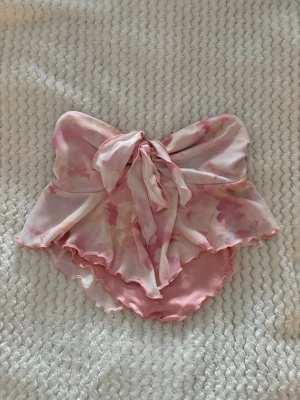 Söt bandeau baddräkt med rosett - Superfin bandeau baddräkt i ljusrosa med blommigt mönster och volangdetaljer. Baddräkten har en stor rosett framtill och är axelbandslös, vilket ger en romantisk vibe. Materialet är mjukt och stretchigt, perfekt för strand eller pool.