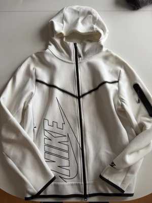 Vit Nike tech fleece - Snygg vit hoodie från Nike med svart dragkedja och stora svarta logotypdetaljer framtill. Hoodien har huva, långa ärmar och svarta detaljer vid ärmslut och dragkedja. Perfekt för en sportig och trendig look.