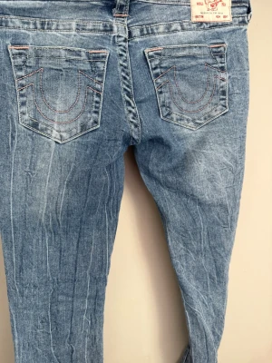 Blå bootcut jeans med kontrastsömmar - Säljer ett par ljusblå bootcut jeans med snygga kontrastsömmar på bakfickorna och slitna detaljer. Jeansen har klassisk femficksdesign och är tillverkade i denim med lätt stretch. Perfekta för en avslappnad och trendig look. Har slitningar längst ner ( se sista bild)