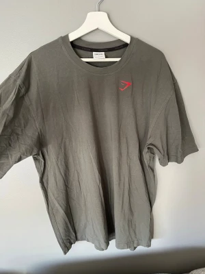 Grå Gymshark t-shirt i bomull - Snygg grå t-shirt från Gymshark med diskret logga i rött på bröstet. Modellen är klassisk med rund hals och korta ärmar, perfekt för en avslappnad stil. Tillverkad i mjuk bomull som känns skön mot huden.