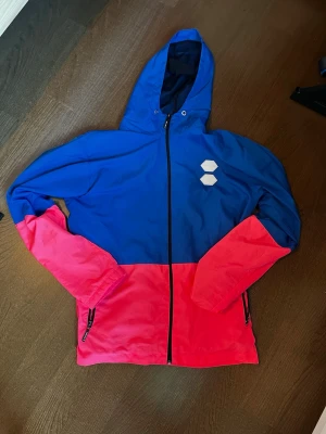  Off-White jacka - Snygg vindjacka från Off-White i blått och rosa med två vita hexagonpatchar på bröstet. Jackan har huva med dragsko, hel dragkedja framtill och svarta dragkedjor på fickorna. Perfekt för dig som vill sticka ut med färg och streetwear-vibe.