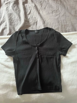 Svart ribbad topp med knappar - En svart ribbad topp från Gina Tricot med korta ärmar och rundad halsringning. Toppen har tre dekorativa knappar framtill och är croppad i modellen. Perfekt att styla med jeans eller kjol för en enkel och snygg look. Storleken är M men är ganska liten i storleken, mer som en S.