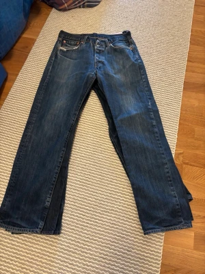 Blå raka jeans från Levi's - Säljer ett par klassiska blå jeans från Levi's med rak passform och fem fickor. Jeansen har en mörk tvätt med lätt slitning och är tillverkade i kraftig denim. Perfekta för dig som gillar en tidlös och avslappnad stil.