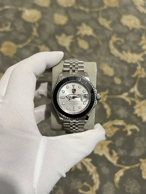 Seiko GMT (PORCHE) Automatisk - Stilren klocka i silver med länkarmband och vit urtavla. Klockan har en svart vridbar ring runt urtavlan och tydliga indexmarkeringar. Mekaniskt urverk syns genom baksidan. Passar dig som gillar klassisk och tidlös design.