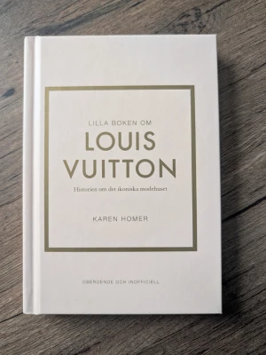 Lilla boken om Louis Vuitton - En stilren bok med vit hård pärm och guldfärgad text. Boken handlar om historien bakom det ikoniska modehuset Louis Vuitton och är skriven av Karen Homer. Perfekt för dig som gillar mode och design.